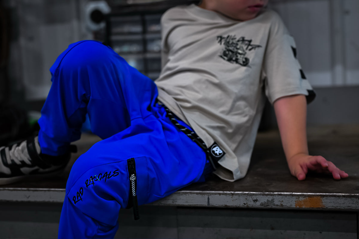 A person sitting on a metal ledge wearing bright blue streetwear pants with black graphics and a beige graphic t-shirt, in a dimly lit industrial setting.