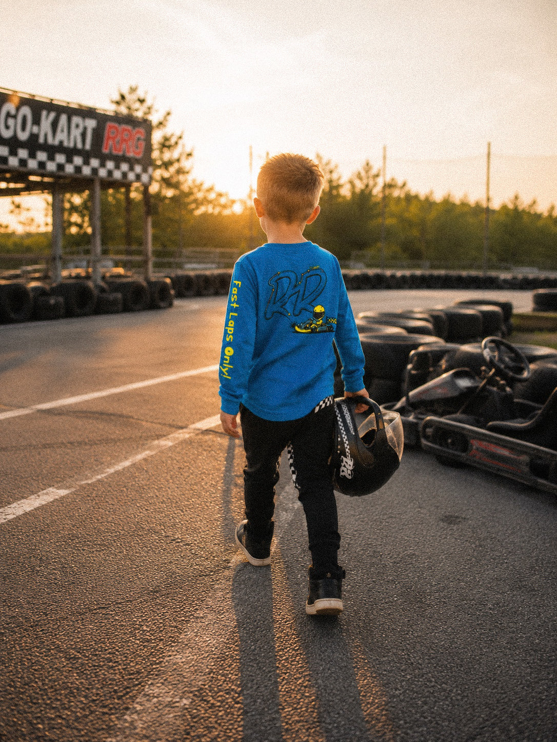 A young boy wearing a bright blue long-sleeve go-karting shirt and black pants walks away on an asphalt track at sunset, holding a helmet. Go-karts and a checkered sign are visible in the background.