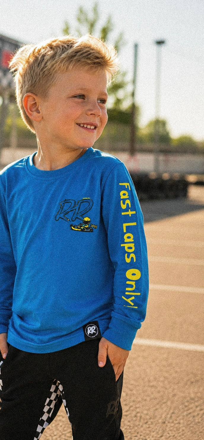 A young boy wearing a bright blue long-sleeve t-shirt with 'fast Laps only!' printed in yellow down the sleeve, standing outdoors on a sunny day.