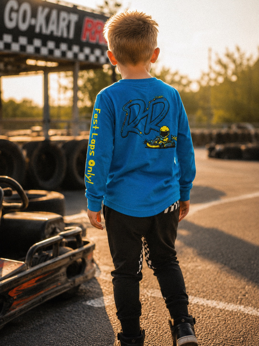 A young boy stands on an outdoor go-kart track wearing a bright blue long-sleeve shirt with a large graphic on the back and black pants, photographed from behind in warm golden hour light.