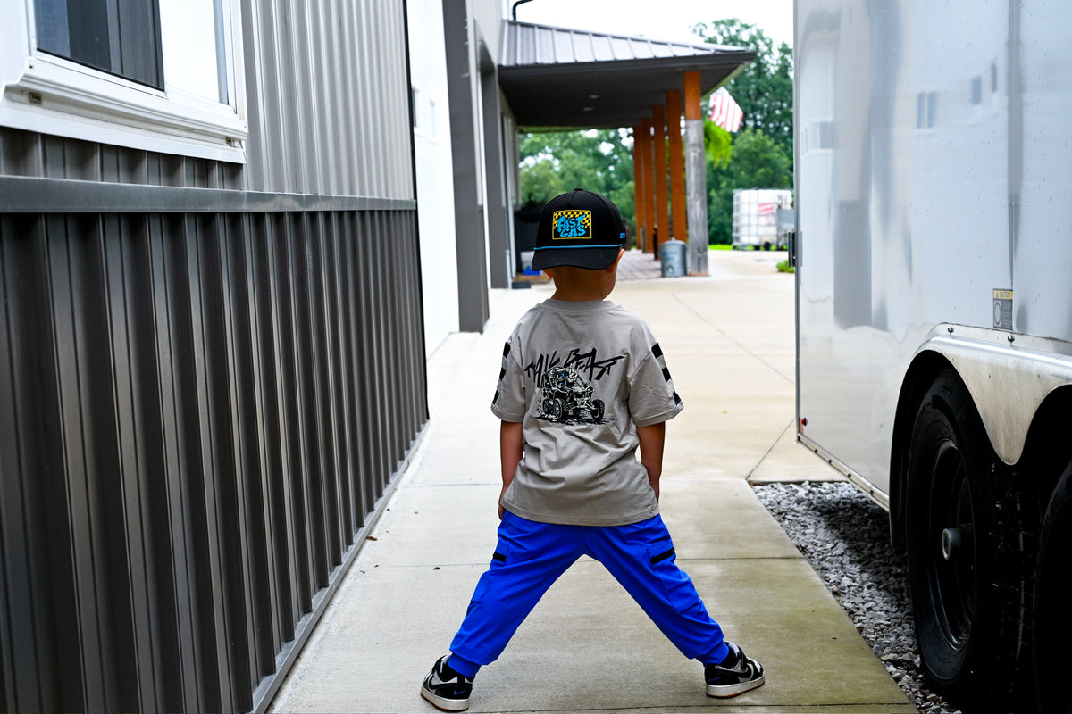 A young boy stands in a wide stance on a concrete walkway, wearing a gray graphic t-shirt, bright blue sweatpants, and a black Stussy cap. He is photographed from behind, with a gray corrugated metal building on the left and a large white truck on the right.