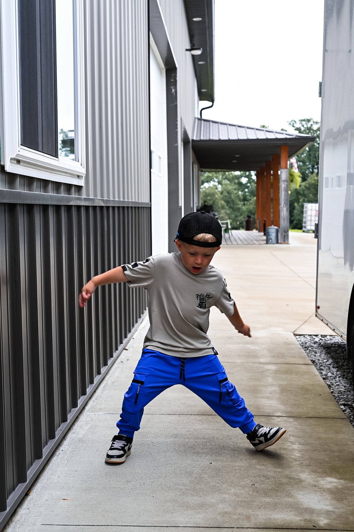 A young boy wearing a gray t-shirt, bright blue cargo pants, and a black cap backwards poses in a dynamic stance on a concrete walkway next to a gray shipping container.
