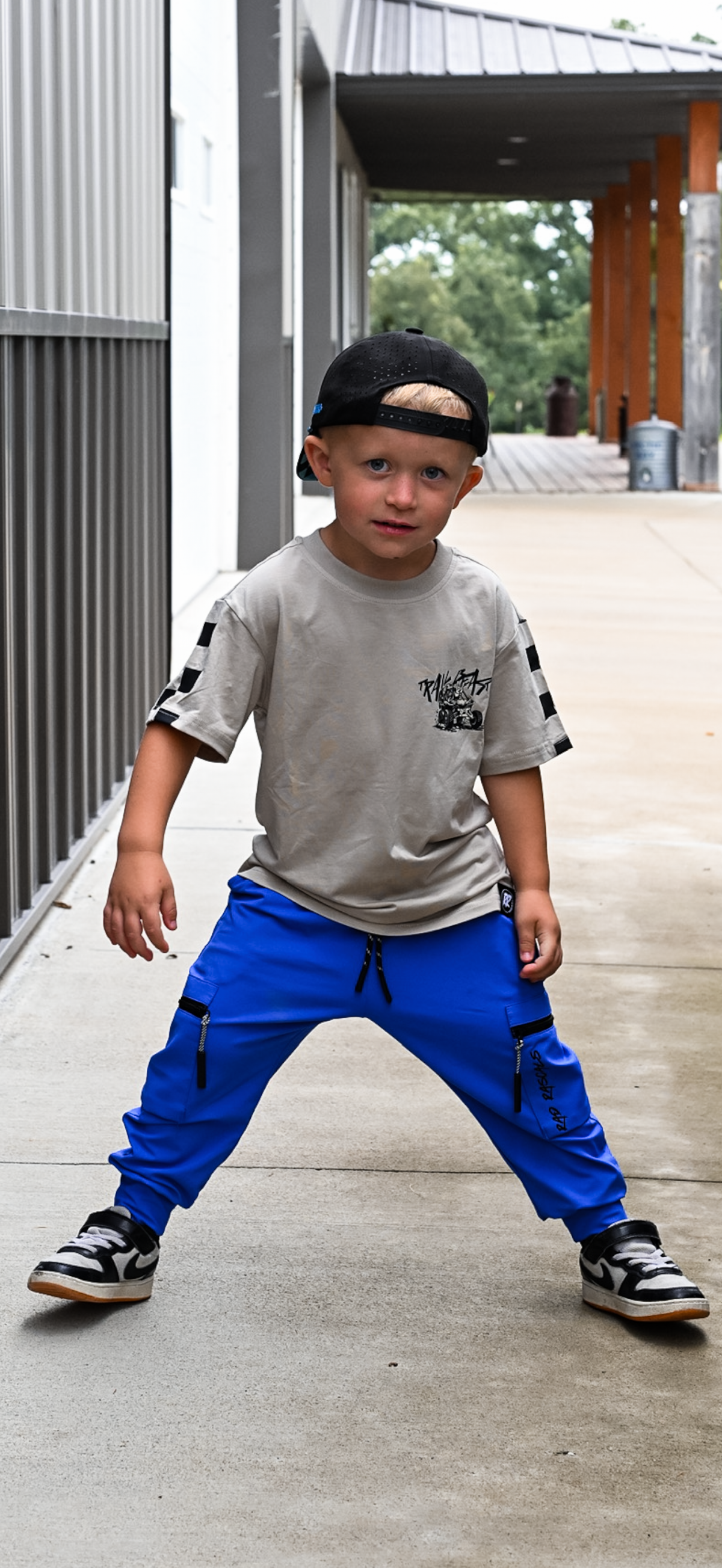 A young boy wearing a gray graphic t-shirt, bright blue cargo pants, and black sneakers poses in a concrete walkway next to a modern building.