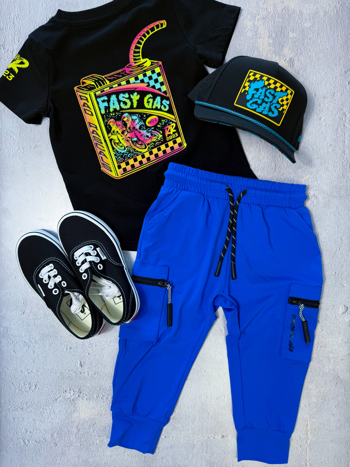 A styled flat-lay outfit set featuring a black graphic t-shirt with a colorful neon 'Easy Gas' design, bright blue jogger pants with black zipper cargo pockets, black sneakers, and a black cap with a matching neon 'Easy Gas' patch.
