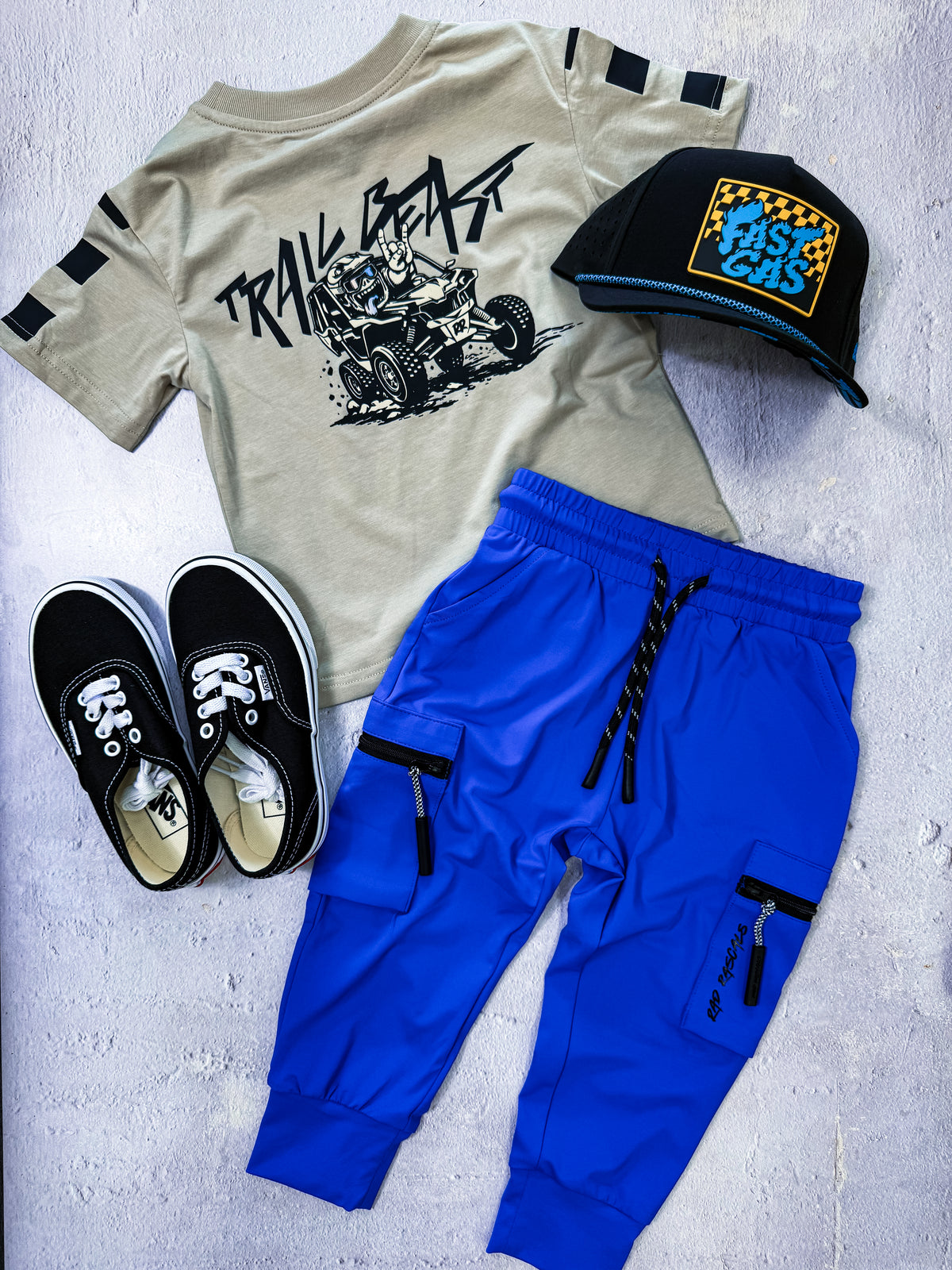 A styled flat-lay outfit set for a child, featuring a beige graphic t-shirt with black sleeve stripes, bright blue jogger pants with black cargo details, black sneakers, and a black cap with a yellow and blue 'Fast Cars' logo, all arranged on a light gray textured background.